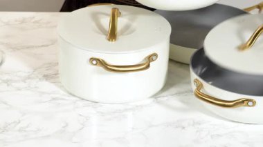 Bir insan beyaz seramik bir tencerenin kapağını açar ve içini ortaya çıkarır. Tencere, parlak ışıklandırmanın altında mermer bir mutfak yüzeyine yerleştirilmiş modern bir yemek takımının parçası..