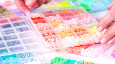 Kadın elleri şeffaf organizatörlerin renklerine göre sıralanmış boncuk koleksiyonunun üzerinde zarif bir şekilde duruyor. Boncuk dizisi, derin morlardan parlak portakallara kadar, titizlikle geniş bir yelpazeyi kapsıyor.