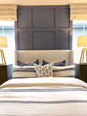 Döşeme yatağı ve arkasında koyu renk bir duvar paneli olan modern bir yatak odası tasarımı. Güneş ışığı boşluğu sıcaklıkla doldururken, lambalar ve komodinlerin eşleşmesi yatağı çerçeveliyor. Atmosfer...