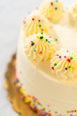 The image shows a tall round cake topped with buttercream swirls, each decorated with multicolored sprinkles. The cake rests on a golden base, and the background is softly out of focus. The