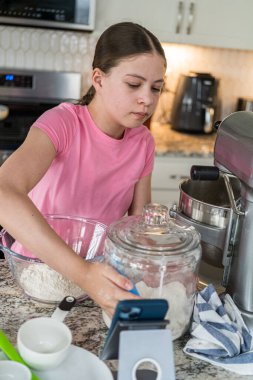Pembe gömlekli bir kız mutfak tezgahında büyük bir un kavanozuna uzanıyor. Ölçme bardakları ve pişirme aletleri yakında düzenlenir.