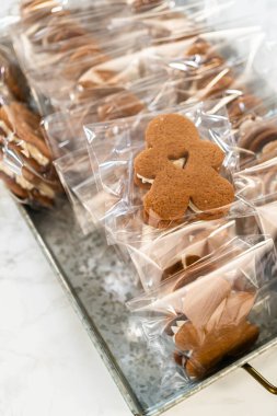 Captured from a close-up perspective, a gingerbread cookie featuring a heart-shaped cutout is individually wrapped in clear packaging, stacked among other holiday treats. The charming design and
