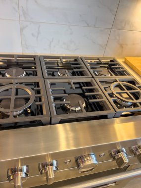 Paslanmaz çelik gaz aralığının ön açılı görüntüsü. Metalik düğmeleri ve fayans arkaplanı var. Görüntü, lüks bir mutfak ortamında son buluyor..