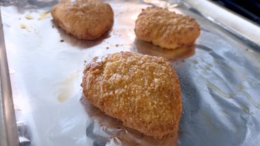 Üç tane kızarmış tavuk göğsü folyo kaplı bir tavada pişiyor. Tavuk, görünür yağlı ve çıtır çıtır bir dış kaplamayla altın kahverengidir..