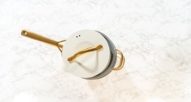 Beyaz kapaklı ve altın saplı tek seramik tencere temiz mermer zemin üzerine kuruludur. Ürün ışıkları, minimalist reklamlar veya öğretici yemek içeriği için uygundur..