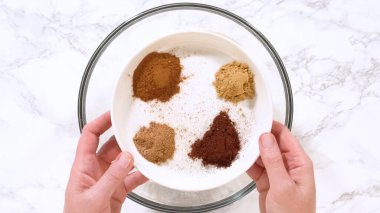 Bir çift el tarçın, hindistan cevizi ve zencefil içeren çeşitli fırın baharatları içeren beyaz bir kase tutuyor. Kase mermer bir tezgahın üzerine yerleştirilmiş. Sahne içeriği vurguluyor