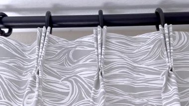 Beyaz soyut dalga desenli gri bir kumaş perdesinin statik yakın çekimi. Perde yumuşak ışıklandırılmış arka planda siyah halkalara asılı..