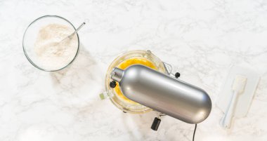 Klasik Şekerli Kurabiye Hamuru malzemeleri içeren elektrikli mikser ve kaselerin üst görüntüsü. Karıştırıcı kısmen karışık malzemelerle bir kasenin üzerine yerleştirilirken, yakındaki kaselerde un ve...