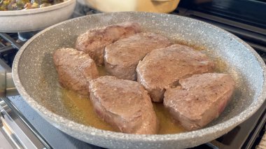 Birkaç biftek madalyonu cızırdayan yağla dolu bir tavada pişiyor. Tava bir gaz sobasının üzerinde duruyor ve altın-kahverengi kabuk düzgün bir kavrulma işareti vermeye başlıyor..