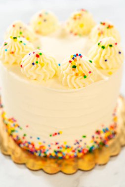 Dikey çerçevelenmiş, üzerinde borulu krema ve renkli yuvarlak serpilmiş uzun beyaz bir pasta olan bir resim. Pasta, yumuşak ışıklandırılmış arka planı olan altın bir tahtada sergileniyor. Genel sahne.