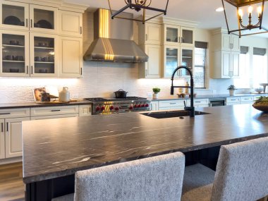 Geniş bir mutfak manzarası geniş bir ada, kolye ışıkları ve paslanmaz çelik aletler. Sahne açık tasarım, parlak ışıklandırma ve zarif ev detaylarını vurguluyor.