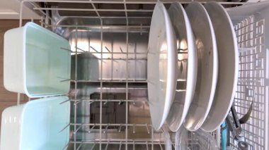 Ağır çekimde A kişisinin eli, bir bulaşık makinesinin açık tepsisinden pırıl pırıl temiz tabak ve çatal bıçak indirirken hareket halinde belirir..