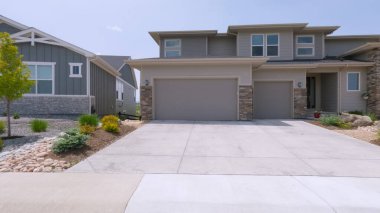 Modern bir banliyö evinde iki arabalık bir garaj, taş süsleme ve minimalist peyzajlı bir ön bahçe bulunur. Açık mavi gökyüzünün altında garaj yolu temiz ve geniştir..