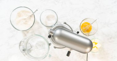 Klasik Şekerli Kurabiye Hamuru malzemeleri içeren elektrikli mikser ve kaselerin üst görüntüsü. Karıştırıcı kısmen karışık malzemelerle bir kasenin üzerine yerleştirilirken, yakındaki kaselerde un ve...