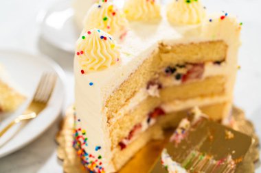 Kısmen dilimlenmiş bir doğum günü pastası altın bir tahtanın üzerinde görünür bir spatula dilimi kaldırıyor. Boru şeklinde krema ve renkli serpinti kenarları süslüyor. Arkaplan levhaları ve çatallar yavaşça tükenmiş.