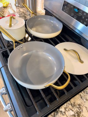 Altın saplı beyaz tencere takımları gaz sobasının tepesine yerleştiriliyor. Temiz mutfak ortamında granit tezgah ve paslanmaz aletler bulunuyor. Parlak ışıklandırma,