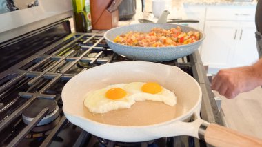 Ev yapımı bir gaz ocağında iki seramik tava var: biri kızarmış yumurta, diğeri sebze ve sosis karışımı..