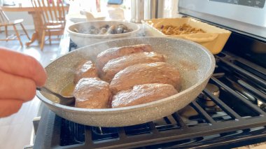Altı çiğ et madalyonu, soba ocağının üzerinde yanmaya başlarken gri bir çubuksuz tavaya konur. Etrafındaki mutfak pırıl pırıl, rahat bir havayla aydınlanıyor..