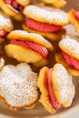 Limonlu spritz kurabiyeli sandviçlerle dolu bir kase, her biri canlı yaban mersini kremalı ve pudra şekerli. Kurabiyelerin narin, tereyağlı ve neşeli bir dokunuşu var.