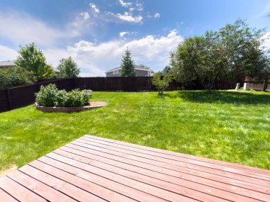 Güneşli bir günde, yeşil çimenlerin, ahşap güvertenin ve yükseltilmiş bahçe yataklarının olduğu geniş bir bahçe. Ağaçlar ve mahremiyet çitleri çimlerin etrafını sarar ve gökyüzünde parlak, dağınık bulutlar vardır..