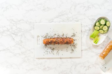 Tütsülenmiş somon ve krem peynirli rulo tamamen susam tohumlarıyla kaplanmış, beyaz bir kesme tahtasına yerleştirilmiş. Taze sebzeler toplanmak için yakınlarda..