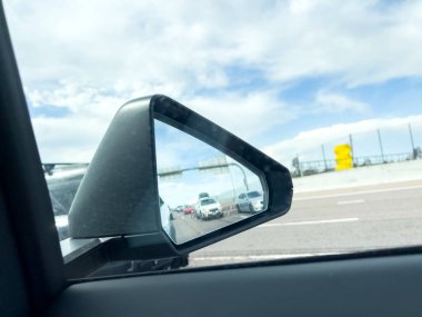 Parker, Colorado, ABD-15 Haziran 2025 - Bir araba yan aynası parçalı bulutlu bir gökyüzünün altında trafik ve yol bariyerlerinin yansımasını yakalıyor. Sahne birden fazla arabası olan bir otoyol ortamı gösteriyor.