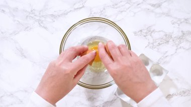 Eller mermer bir tezgaha yerleştirilmiş temiz bir cam karıştırma kabına yumurta kırar. Resim, kurabiye pişirmek için basit bir malzeme hazırlama adımını vurguluyor.