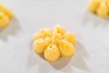 Pişmemiş limonlu spritz kurabiye hamuru parşömen astarlı pişirme tepsisinin üzerinde, kurabiye makinesi kullanarak çiçek deseni şeklinde duruyor. Hamurun tereyağı gibi taze limon tadı var.