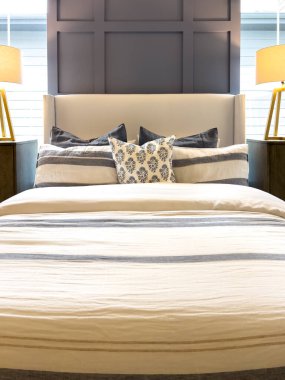 Tarafsız çizgili çarşafları ve dekoratif yastıkları olan rahat bir yatak odası. Alan her iki tarafında da masa lambaları olan komodinleri içeriyor. Yumuşak gün ışığı