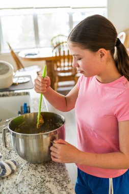 Genç bir kız kurabiye hamurunu granit mutfak tezgahındaki paslanmaz bir kasede spatulayla karıştırıyor. Mutfakta büyük pencerelerden gelen doğal ışığın olduğu bir yemek alanı var..