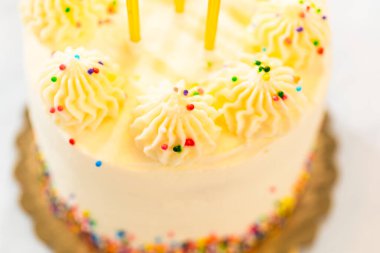 Sarı mumlarla kaplı ve altın rengi bir tabana yerleştirilmiş bir doğum günü pastasının yakından görünüşü. Şekerlemeler küçük renkli süslemelerle süslenmiş ve canlı detaylar eklenmiş..