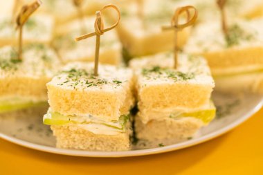 Krem peynir ve salatalık dilimleri ile doldurulmuş, kurutulmuş dereotla kaplı ve bambu kazmalarıyla korunmuş yumuşak beyaz ekmek kareleri. Bu ısırık büyüklüğünde sandviçler beyaz bir tabakta, parlak bir tabakla hazırlandı.