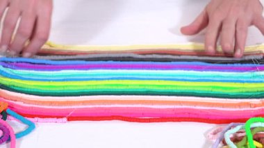 Eller birbirine zıt beyaz bir yüzeye karşı düzgün düzenlenmiş renkli, sarmalanmış kil boncuk iplikçiklerine dokunur..