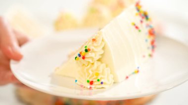 Süslü bir dilim vanilyalı pasta, beyaz krema ve gökkuşağı parçacıkları. Kalan pastanın yanındaki mermer yüzeyden açılı..