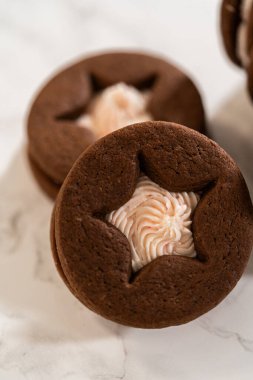 Çikolatalı sandviç kurabiyeleri ve yıldız şekilli şekerlemeler yakın planda gösteriliyor. Yumuşak ışıklandırma yumuşak krema ve zengin kurabiye dokusunu vurgular.