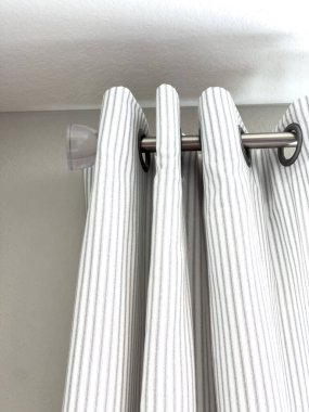 Beyaz çizgili perdeler metal bir çubuğa asılıdır. Kıvrımlı üstleri vardır. Yumuşak ışık girsin diye kısmen açıktır. Odada rahat, nötr renkli halı döşemesi var..