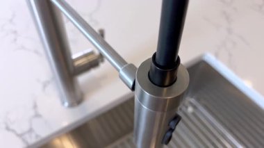 Modern bir lavabonun üzerine yerleştirilmiş paslanmaz çelikten yapılmış bir mutfak musluğunun detaylı görüntüsü. Arka planda yumuşak ışıklandırmada mermer bir tezgah var..