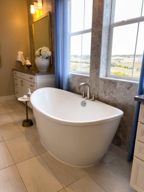 Mavi perdeli bir pencerenin yanında serbest dolaşan beyaz bir küvet içeren zarif bir banyo tasarımı. Tarafsız tonlar ve doğal gün ışığı huzurlu ve ferahlatıcı bir görünüm yaratır. Modern tasarım