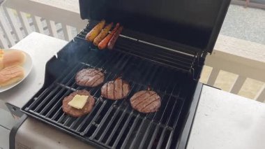 Peynir dilimleri ızgara hamburger köftelerinin üzerine konur. Hamburgerler, ahşap bir verandadaki bir ızgarada sosisin yanında pişerler..