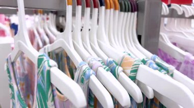 Mağazadaki beyaz plastik askılarda renkli yaz elbiseleriyle dolu bir elbise rafı. Kıyafetlerde sıcak havaya uygun parlak desenler ve hafif kumaşlar var. Arka plan gösteriyor ki...