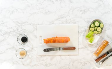 Bıçaklı temiz beyaz bir kesme tahtası, bir blok füme somon, krem peynir, salatalık kurdeleleri, ve siyah-beyaz susam tohumları, hazırlanmaya hazır..