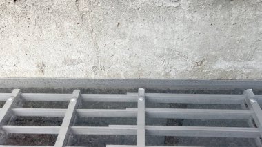 Beton bir temele monte edilmiş gri metal bir pencere ızgarasının yakın çekimi. Resim çubukların ve yüzey temaslarının hizalanmasını vurgular.