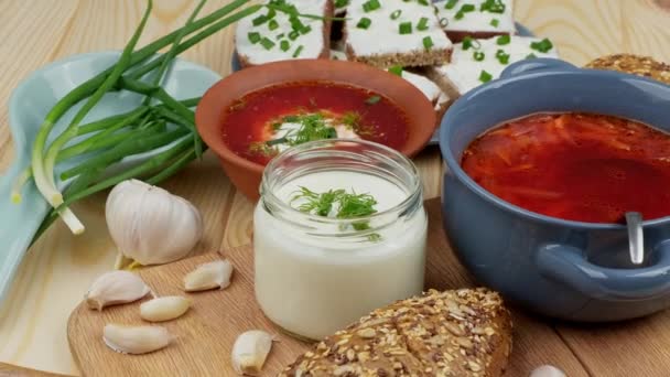 Russian and Ukrainian cuisine. Borscht with sour cream and spices, fresh herbs, garlic and pampushki. Beetroot soup in a blue bowl on a wooden board. — Stock Video