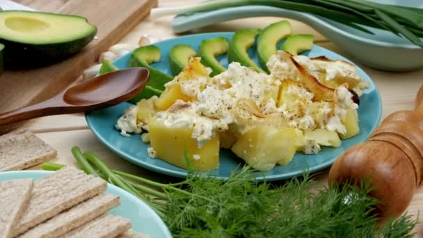 Pečené brambory se sýrem s plátky avokáda a čerstvými bylinkami na modrém talíři na dřevěném stole — Stock video