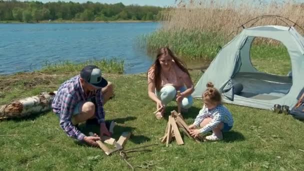 Семья отдыхает на берегу реки с палаткой. Мать и дочь собирают дрова — стоковое видео