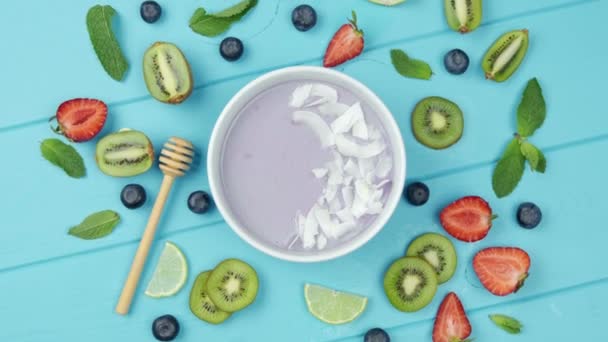 Tazón de batido con arándanos y trozos de coco, decorado con fruta fresca. concepto de comida saludable. Fondo de madera azul — Vídeos de Stock