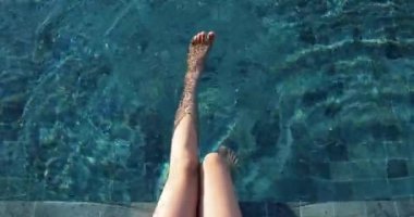 Yazın güneşin altında, tatil köyü havuzundaki mavi suya karşı ince bacakların yakın çekimi. Kadın bacaklarını havuzun içinde sallıyor. Resimleri kavramsallaştır, rahatla, seyahat et, tatil yap. 4K.