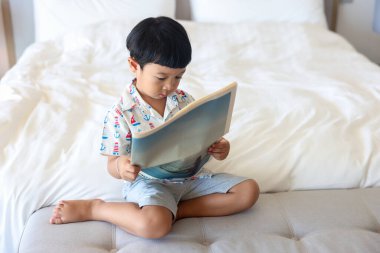 Kâhküllü, neşeli saçlı Asyalı bir çocuğun portresi yatakta oturup, beyaz gömlek giyen, aktif 4-5 yaşındaki bir çocuğun komik bir şekilde gülümseyen ve kameraya bakan sevimli bir çocuk..