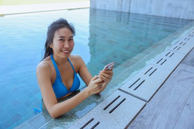 Güzel Asyalı genç kadın portresi, mavi bikinili otelin havuzunda kameraya gülümseyen akıllı telefon kullanıyor. Arka planda mavi yüzme havuzu.