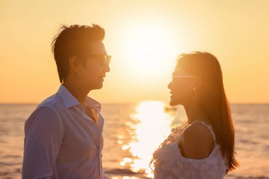 Genç mutlu Asyalı çift sahilde birbirine bakıyor, romantik seyahat balayı tatili yaz tatili. Asyalı kadın ve erkek, gün batımında el ele tutuşuyorlar.